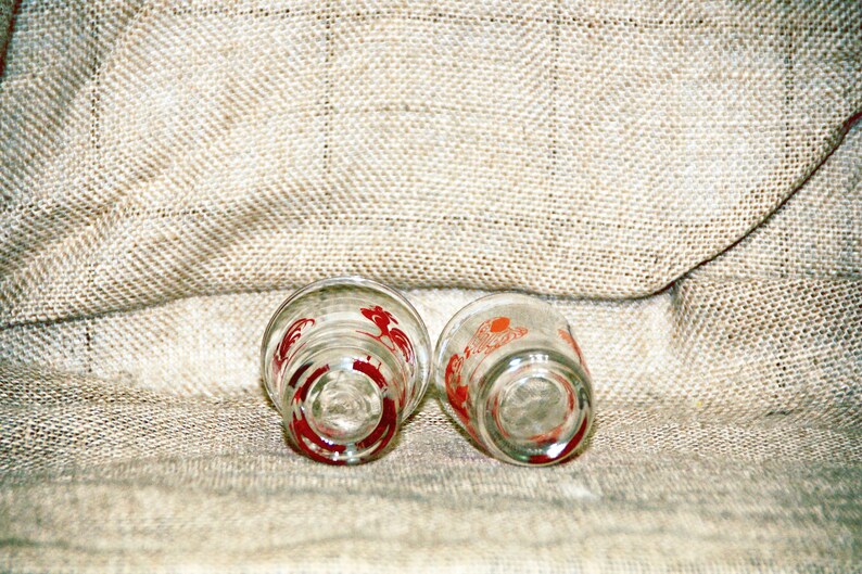 May include: Two clear glass shot glasses with red rooster designs. The glasses are upside down on a burlap surface.