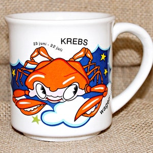 May include: White ceramic mug with a cartoon crab illustration. The crab is orange and has a blue background with stars. The text "KREBS 23 JUNI - 22 JULI" is printed on the mug.