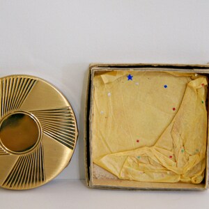 May include: A gold compact with a sunburst design, in its original box. The box is made of cardboard and has a yellow tissue paper lining.