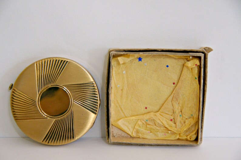 May include: A gold compact with a sunburst design, in its original box. The box is made of cardboard and has a yellow tissue paper lining.