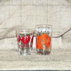 May include: Two clear glass drinking glasses with red and orange designs. The smaller glass has a red rooster design and the taller glass has an orange rooster design.