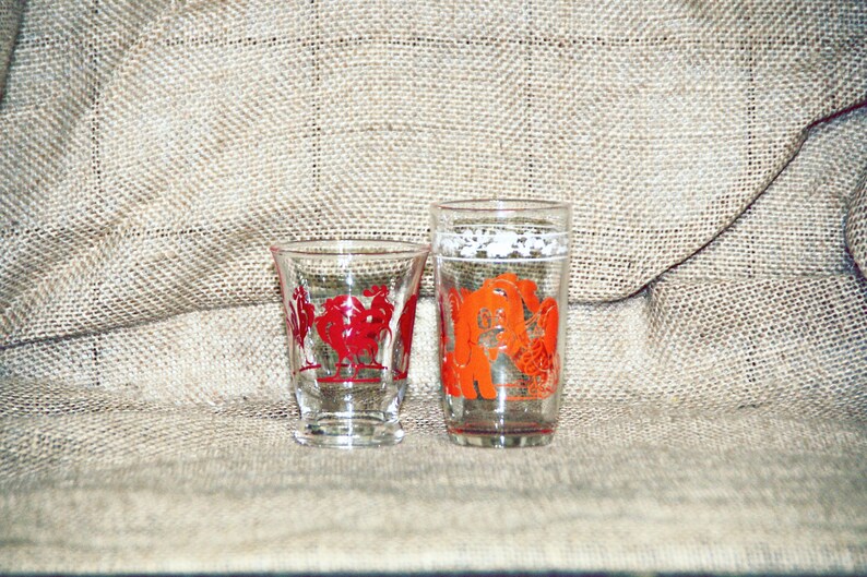 May include: Two clear glass drinking glasses with red and orange designs. The smaller glass has a red rooster design and the taller glass has an orange rooster design.