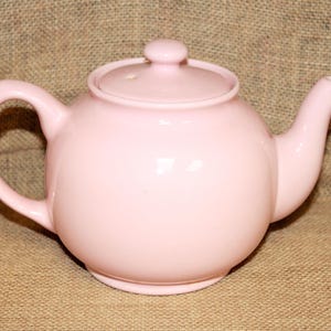 May include: A pale pink ceramic teapot with a rounded body and a lid. The teapot has a spout and a handle.