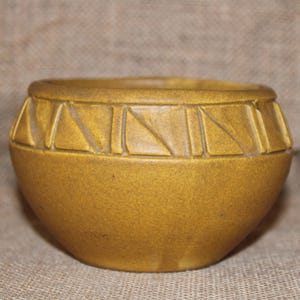 May include: A round, mustard-yellow ceramic bowl with a textured surface. The rim features a repeating geometric design of triangles. The bowl is a decorative item, suitable for holding small objects or as a display piece.