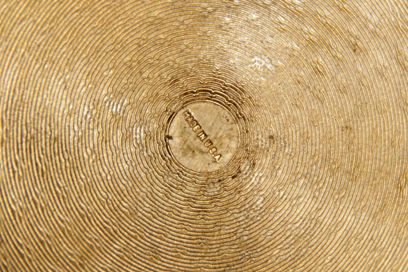 May include: Close-up of a gold-colored metal surface with concentric circles. The center of the surface has a small, circular stamp that reads "Made in U.S.A."