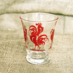 May include: A clear glass drinking glass with a red rooster design. The rooster is in a standing position with its wings spread out.