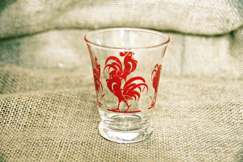 May include: A clear glass drinking glass with a red rooster design. The rooster is in a standing position with its wings spread out.