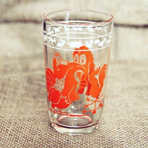 May include: A clear glass drinking glass with a red rooster and a compass design. The glass has a white border with a repeating pattern of animals.
