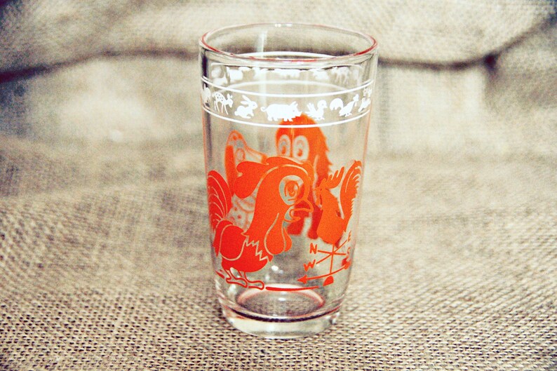 May include: A clear glass drinking glass with a red rooster and a compass design. The glass has a white border with a repeating pattern of animals.