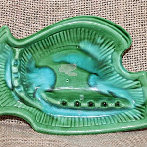 May include: Green ceramic ashtray shaped like a fish with a blue glaze. The ashtray has a raised rim and a series of holes for cigarettes.