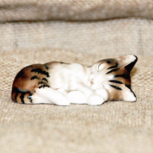 May include: A small, ceramic figurine of a sleeping tabby cat. The cat is curled up on its side with its paws tucked under its chin. The figurine is painted in shades of brown, white, and black.