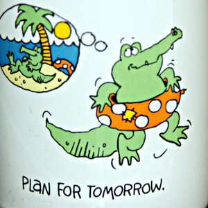 May include: White ceramic mug with a green alligator wearing an orange and white life preserver. The alligator is swimming in a blue body of water with a palm tree and the text "Plan for tomorrow."
