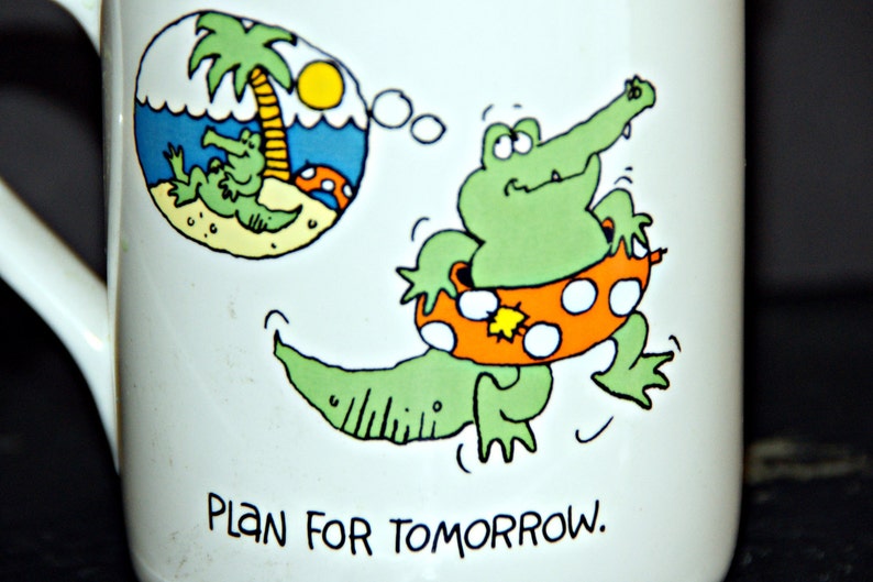 May include: White ceramic mug with a green alligator wearing an orange and white life preserver. The alligator is swimming in a blue body of water with a palm tree and the text "Plan for tomorrow."