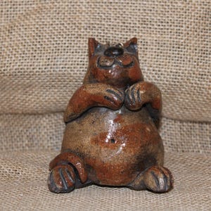 May include: A ceramic cat figurine in a seated position, with paws clasped together. The cat has a brown and tan color scheme, with a glossy finish. The figurine is a whimsical decorative item.