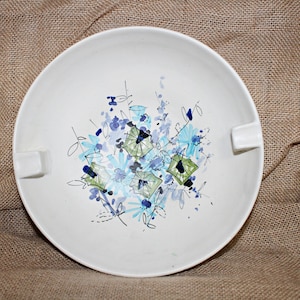 May include: A white ceramic bowl with a blue and green floral design. The bowl has a lip on one side and is perfect for serving snacks or dips.