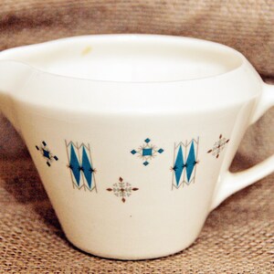 May include: White ceramic creamer with a blue and brown geometric pattern. The creamer has a handle and a spout.