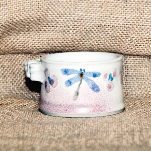 Könnte beinhalten: Eine kleine weiße Keramiktasse mit zwei Henkeln, verziert mit einem handgemalten Design einer blauen Libelle und rosa Blumen.
