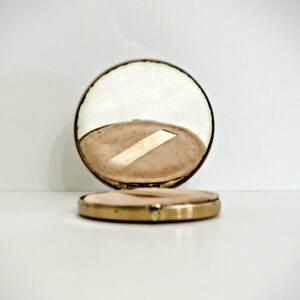 May include: A gold compact mirror with a round, mirrored lid and a white interior. The lid is open, revealing a white, padded interior.