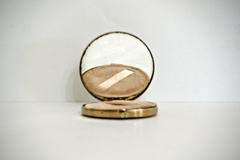 May include: A gold compact mirror with a round, mirrored lid and a white interior. The lid is open, revealing a white, padded interior.
