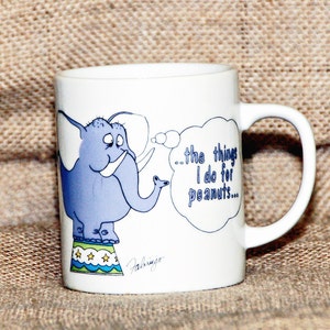 Könnte beinhalten: Weiße Keramiktasse mit einer Cartoon-Illustration eines blauen Elefanten, der auf einer bunten Plattform steht. Der Elefant denkt in einer Gedankenblase: "...die Dinge, die ich für Erdnüsse tue..."