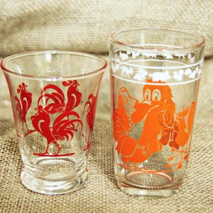 May include: Two clear glass drinking glasses with rooster designs. The smaller glass has red roosters and the larger glass has an orange rooster.