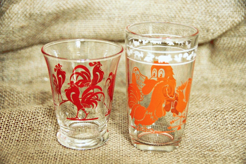May include: Two clear glass drinking glasses with rooster designs. The smaller glass has red roosters and the larger glass has an orange rooster.