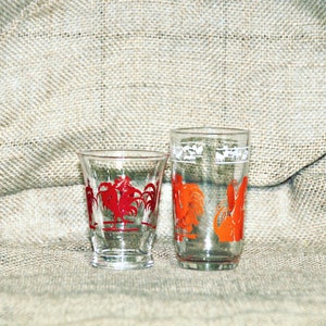 May include: Two clear glass drinking glasses with rooster designs. The smaller glass has a red rooster design and the taller glass has an orange rooster design.