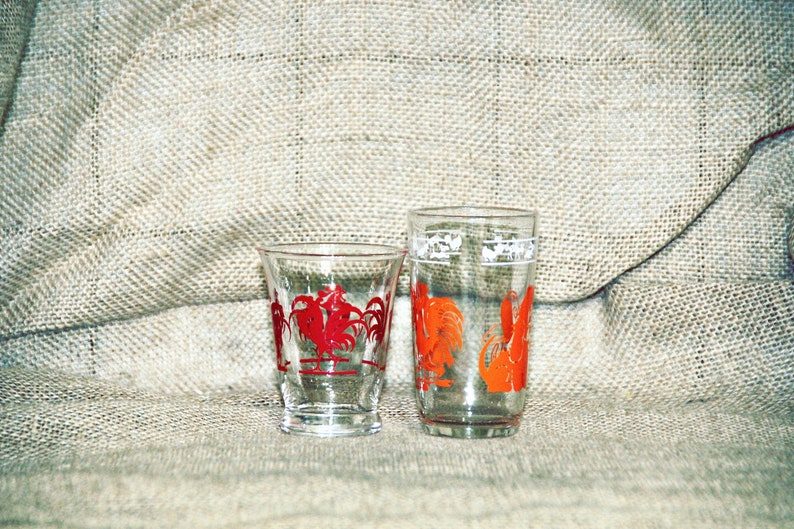 May include: Two clear glass drinking glasses with rooster designs. The smaller glass has a red rooster design and the taller glass has an orange rooster design.