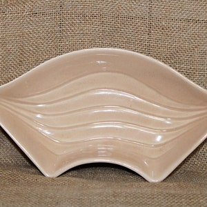 Op de afbeelding: Een beige keramische schaal met een unieke, waaiervormige vorm. De schaal heeft een reeks gebogen, golfachtige ribbels over het oppervlak. Het ontwerp is eenvoudig en elegant, geschikt om te serveren of te presenteren.