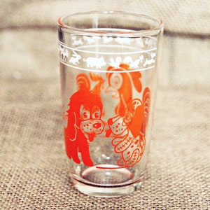 May include: A clear glass drinking glass with a red and white design of two dogs. The dogs are outlined in white and filled in with red. The glass has a white band around the top with a design of animals.