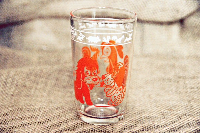 May include: A clear glass drinking glass with a red and white design of two dogs. The dogs are outlined in white and filled in with red. The glass has a white band around the top with a design of animals.