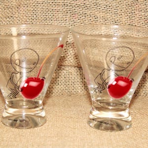 May include: Two clear martini glasses with a quirky design. Each glass has a cartoon figure holding a bright red cherry. The glasses are set against a neutral background, highlighting the playful design.