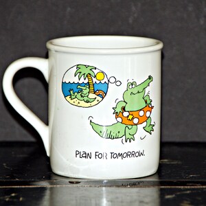 May include: White ceramic mug with a green alligator wearing a life preserver and a thought bubble with a cartoon of the alligator relaxing on a beach. The mug reads "Plan for Tomorrow."