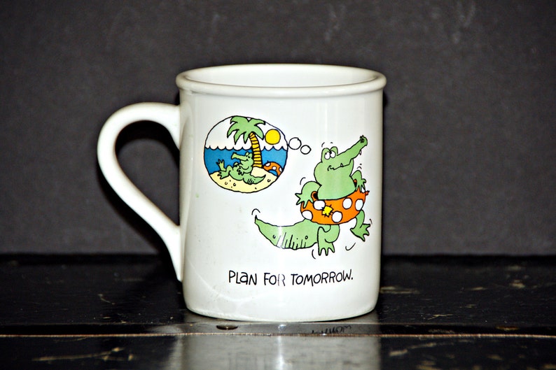 May include: White ceramic mug with a green alligator wearing a life preserver and a thought bubble with a cartoon of the alligator relaxing on a beach. The mug reads "Plan for Tomorrow."