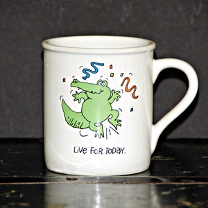 May include: White ceramic mug with a green alligator cartoon illustration and the text "Live For Today."
