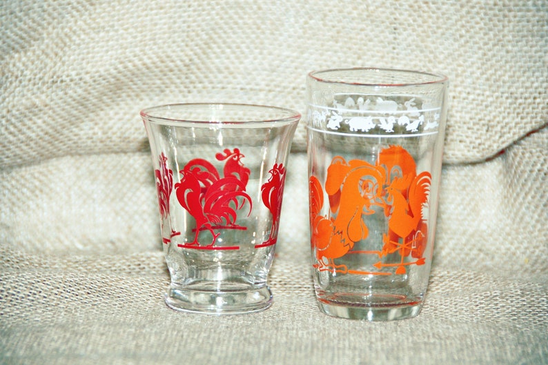 May include: Two clear glass drinking glasses with rooster designs. The smaller glass has red roosters and the taller glass has orange roosters.