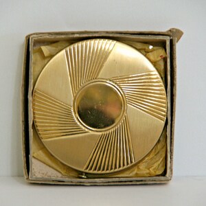 May include: A gold-colored compact mirror with a sunburst design. The mirror is in a cardboard box.