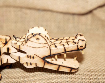 May include: A wooden alligator sculpture with detailed pyrography. The alligator's head is open, revealing teeth. The wood has a light brown color with darker brown burn marks outlining the features. The sculpture is set against a neutral background.