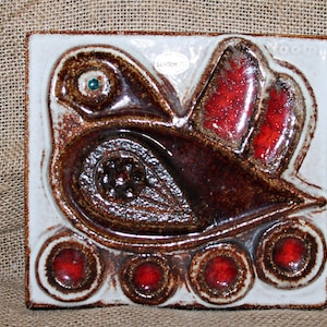 May include: A square ceramic wall tile featuring a relief design of a bird in shades of dark brown and vibrant red. The bird has a small teal eye, and the bottom edge is decorated with four circular red accents against a white background.