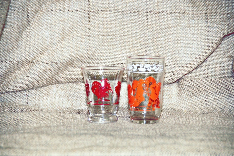 May include: Two clear glass drinking glasses with red and orange rooster designs. The glasses are on a burlap background.