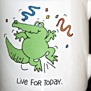 May include: White ceramic mug with a green alligator cartoon illustration and the text "Live for today".
