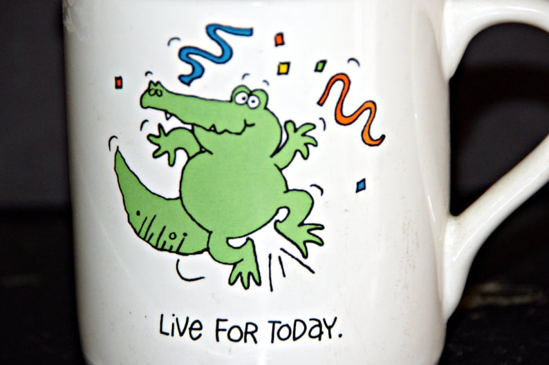 May include: White ceramic mug with a green alligator cartoon illustration and the text "Live for today".