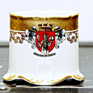 May include: White ceramic cup with a gold rim and a coat of arms design. The coat of arms features a red shield with a gold crown, a man holding a key and a book, and the text "Bremervorde".