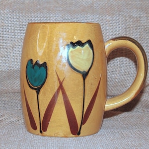 May include: A yellow ceramic mug with a rounded shape and a handle. The mug is decorated with two tulip flower designs in teal and yellow, with brown stems and leaves. The mug is likely handmade.