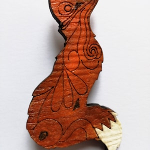 May include: Wooden fox-shaped ornament with intricate etched details. The fox is primarily orange-brown with a lighter, natural wood tail. The design features swirling patterns and leaf-like shapes, creating a rustic, handcrafted aesthetic.