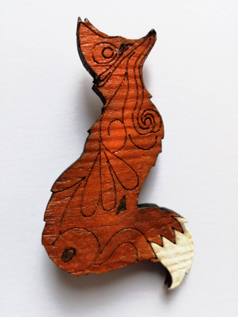 May include: Wooden fox-shaped ornament with intricate etched details. The fox is primarily orange-brown with a lighter, natural wood tail. The design features swirling patterns and leaf-like shapes, creating a rustic, handcrafted aesthetic.