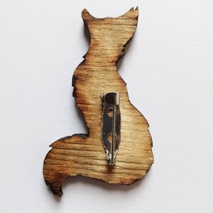 May include: Wooden fox-shaped brooch with a silver pin backing. The brooch is made of light brown wood with darker brown edges, giving it a rustic appearance. The fox silhouette is detailed, showing the ears, body, and tail.