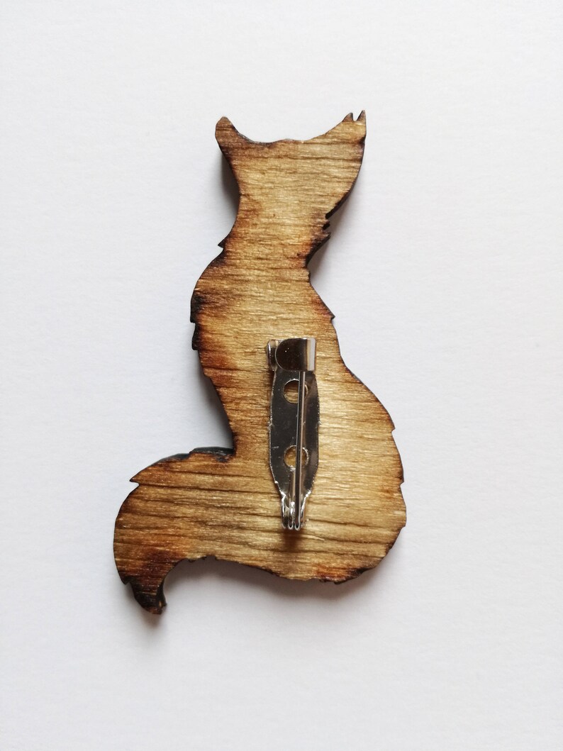 May include: Wooden fox-shaped brooch with a silver pin backing. The brooch is made of light brown wood with darker brown edges, giving it a rustic appearance. The fox silhouette is detailed, showing the ears, body, and tail.
