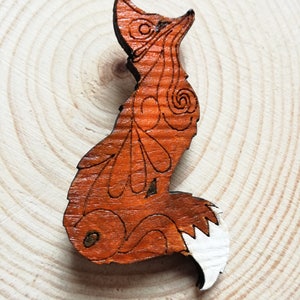 May include: A wooden fox-shaped brooch with intricate etched details. The brooch features a reddish-brown body with a white-tipped tail. The fox is posed in a seated position, with its head turned upwards. The brooch is set against a light wood background.
