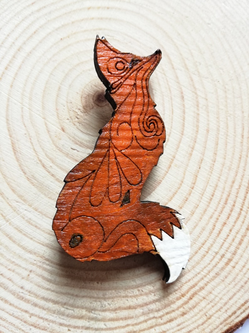 May include: A wooden fox-shaped brooch with intricate etched details. The brooch features a reddish-brown body with a white-tipped tail. The fox is posed in a seated position, with its head turned upwards. The brooch is set against a light wood background.